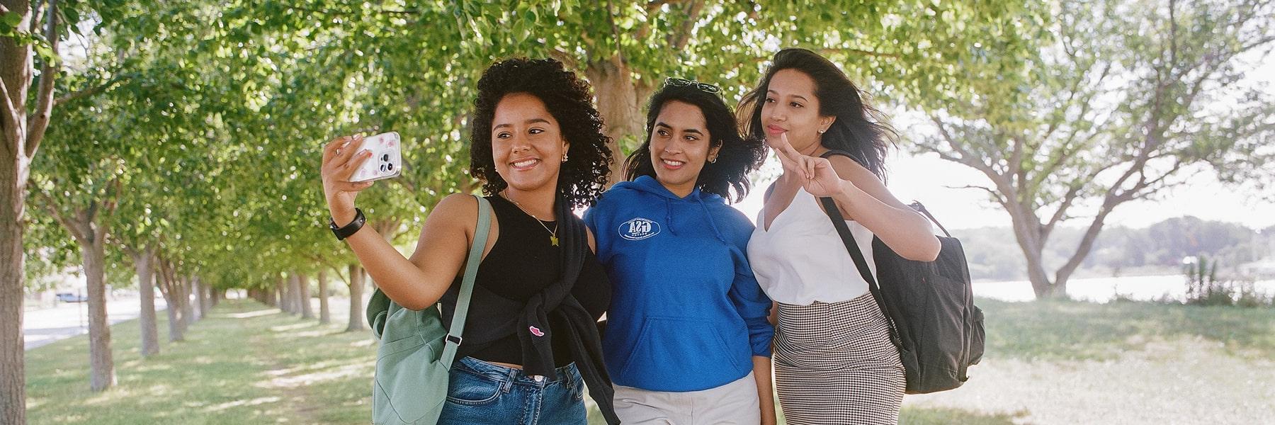 3 students take selfie under trees.